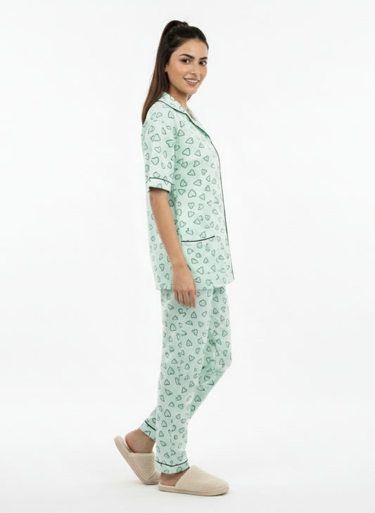 Effortlessly cute and comfortable Koozy pajama set in a refreshing light mint green with a white heart print. A Pinterest-style collared nightwear suite made from breathable cotton fabric, featuring a short-sleeve button-down top with dark green contrast piping and matching long pants. Relaxed fit, beautifully printed PJ set for a soft, easy, and cozy look, styled with plush beige slippers.