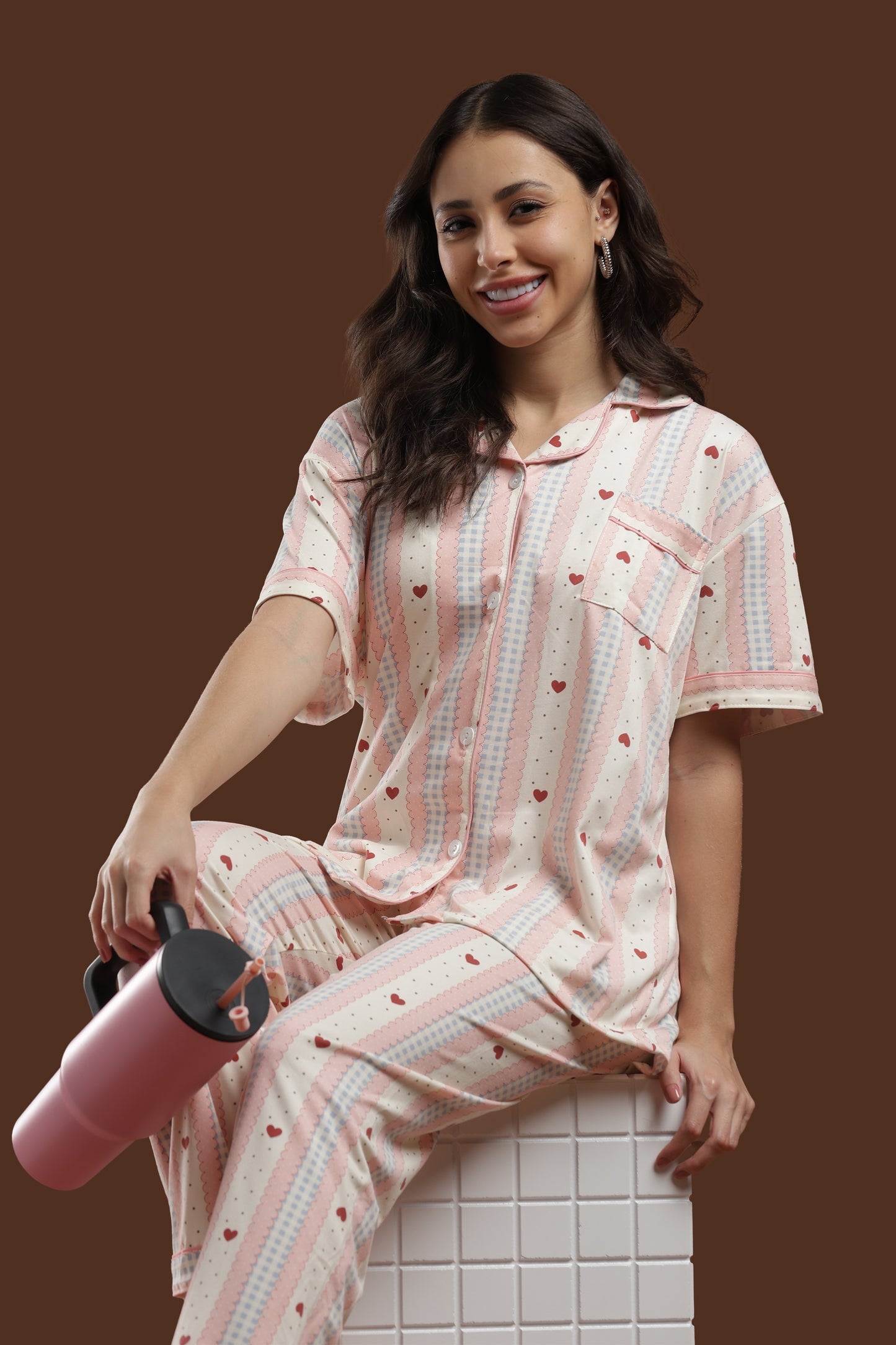 a woman posing in the koozy pink and white colour, with red heart pj set with a pink tumbler, styled for a pinteresty morning vibe. highlights the cute, effortless look of the button-down top and matching pants.